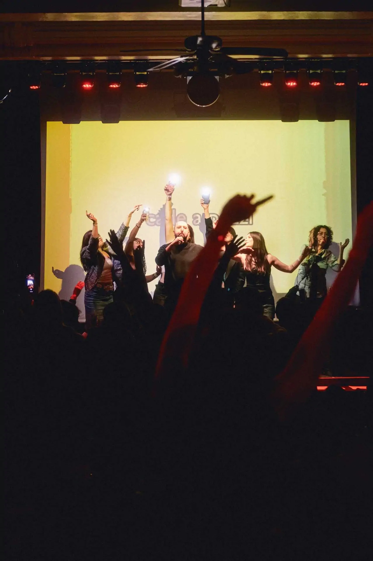 People chanting in front of a crowd, with a projected light behind, in a dark theater.